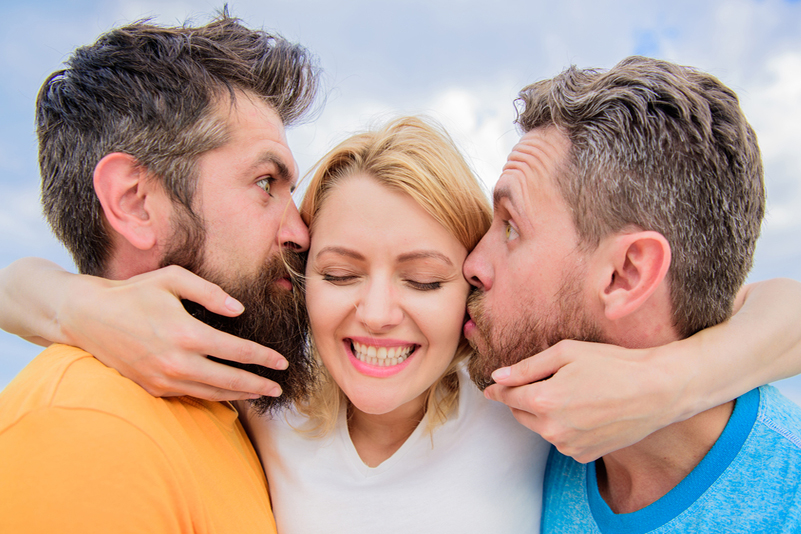 A smiling woman stands between two men with her arm around both their necks. They look confused or agitated.
